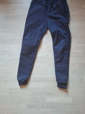 Mörkblå mjukisbyxor med muddar - Säljer ett par mörkblå mjukisbyxor med resår i midjan och snörning. Byxorna har två sidofickor och ribbade muddar vid bensluten. Perfekta för chill dagar eller när du vill vara extra bekväm.