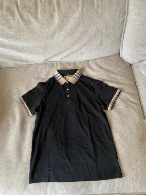 pike - Snygg svart pikétröja med klassisk krage och knappar framtill. Kragen och ärmsluten har beige, röda och svarta ränder i rutigt mönster som ger en stilren kontrast. Perfekt till jeans eller shorts för en avslappnad look.