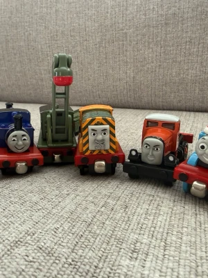 Tågfigurer från Thomas & Vännerna - Säljer ett set med fem färgglada tågfigurer från Thomas & Vännerna. Figurerna är i plast och metall och har olika ansikten och färger som blå, grön, orange och röd. Perfekt för dig som gillar att samla eller leka med tåg och klassiska karaktärer.