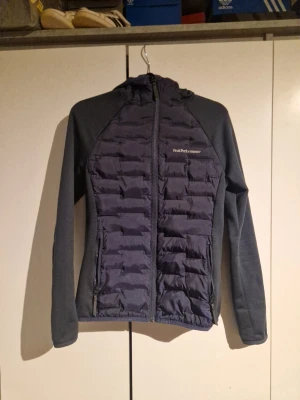 Peak performance jacka - Snygg mörkblå vindjacka från Peak Performance med quiltad front och rygg, samt mjuka ärmar och sidor i stretchigt material. Jackan har hel dragkedja, två fickor med dragkedja och en skön huva. Perfekt för blåsiga dagar och outdoor-aktiviteter.