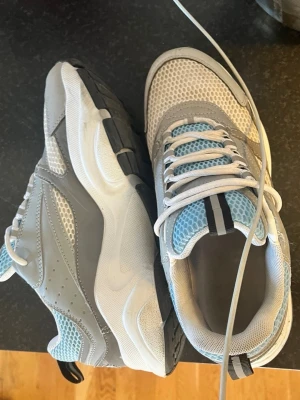 B22 - Säljer ett par chunky sneakers grått, vitt och ljusblått. Skorna har meshpaneler, vita skosnören och en robust sula med svarta och blå detaljer. Perfekta för dig som gillar en sportig och trendig look.