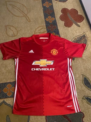 Manchester United matchtröja Pogba - Säljer en röd Manchester United matchtröja från Adidas med Pogba och nummer 6 på ryggen. Tröjan har vita detaljer, klubbmärke på bröstet och Chevrolet-logga framtill. Tillverkad i lätt och ventilerande material, perfekt för fotboll eller träning.STL L