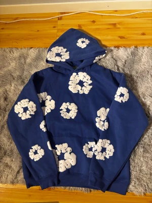 Blå hoodie med vita blommor Denim Tears - Snygg blå hoodie från Denim Tears med vita blommönster över hela tröjan och huvan. Hoodien har en klassisk känguruficka och ribbade muddar. Perfekt för dig som vill sticka ut med en unik och trendig stil.