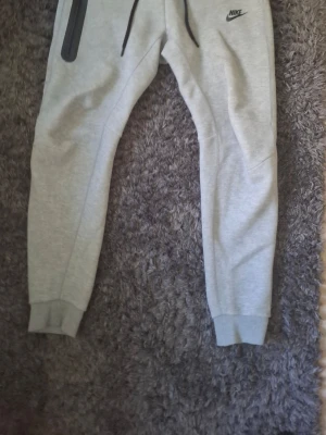 Gråa Nike mjukisbyxor med dragkedja - Snygga gråa mjukisbyxor från Nike med svarta detaljer och dragsko i midjan. Byxorna har en dragkedjeförsedd ficka på ena benet och ribbade muddar vid bensluten. Perfekta för chill eller träning.