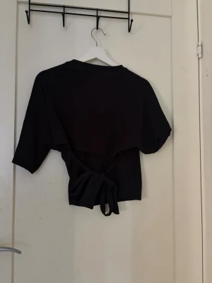 Svart topp med knyt i midjan - Säljer en svart topp med korta ärmar och knyt i midjan. Toppen har en enkel, stilren design och är perfekt för att skapa en avslappnad men ändå snygg look. Materialet känns mjukt och bekvämt mot huden.