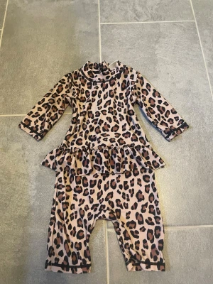 Leopardmönstrad uvdräkt  - Säljer en långärmad stranddräkt för barn med leopardmönster i beige, brunt och svart. Dräkten har volang i midjan, dragkedja bak och långa ben. Perfekt för soliga dagar på stranden och ger extra skydd mot solen.