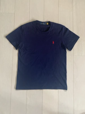 Marinblå t-shirt från Polo Ralph Lauren - Klassisk marinblå t-shirt från Polo Ralph Lauren med röd broderad logga på bröstet. T-shirten har rund halsringning och korta ärmar. Tillverkad i mjuk bomull för en skön känsla och perfekt passform.