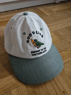 Linne keps med broderad fågel - Säljer en stilren linne keps med vit krona och grön skärm. Framtill finns ett broderat motiv av en grön fågel och texten 'GOOD LIDS' samt 'ENDEMIC TO THE SOUTHERN ALPS'. Kepsen har broderade ventilationshål och justerbar passform. Tillverkad i linne