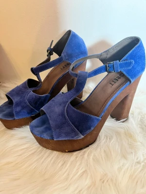 Scorett platåsandaler med träklack - Skor från Scorett. Säljer ett par snygga blå sandaler med breda remmar i sammet och kraftig träklack. Skorna har öppen tå, justerbart spänne vid ankeln och en cool retrovibe. Perfekta för dig som vill sticka ut med färg och höjd.