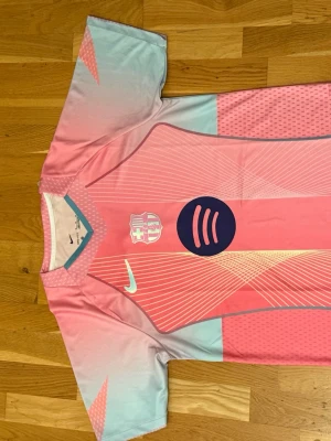 Barça x Nike rosa matchtröja - Säljer en Barcelona matchtröja från Nike i rosa och ljusblått med klubbmärke och Spotify-logga på bröstet. Tröjan har Dri-FIT-material som andas och är perfekt för fotboll eller träning. Snygga grafiska linjer och korta ärmar ger en modern look.