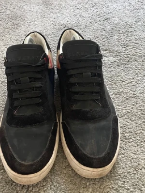Svarta Burberry sneakers med mockadetaljer - Snygga svarta sneakers från Burberry med ovandel i mocka och textil. Klassisk låg modell med svarta skosnören och vit sula. Sidan har Burberrys ikoniska rutmönster i beige, svart och rött. Perfekta för dig som vill ha en stilren och trendig look. Riktigt sköna skor men säljer nu när jag tröttnat på dem, vid fler frågor eller funderingar tveka inte på o skriva! Pris kan alltid diskuteras.