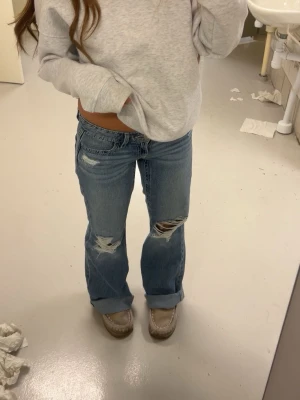 Blå slitna bootcut jeans gina tricot - Säljer ett par ljusblå bootcut jeans med slitningar och hål på båda benen. Jeansen har låg midja och är uppvikta nertill för en avslappnad vibe. Perfekta för dig som gillar en trendig och casual stil. Använd 2 gånger