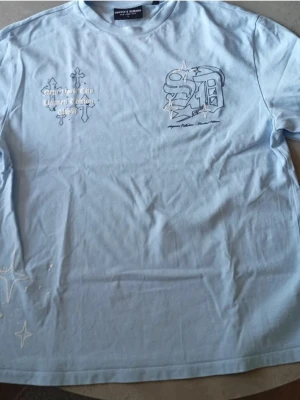 Ljusblå Supply and Demand t-shirt - Snygg ljusblå t-shirt med coola tryck på både fram- och baksidan. Framtill finns grafiska detaljer och text, medan baksidan har ett stort 'supply'-tryck i svart. Tillverkad i mjuk bomull och har en avslappnad passform med korta ärmar.