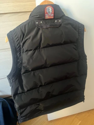 Parajumper Kobuk Väst - Snygg svart kobuk från Parajumpers med hög krage, dragkedja och tryckknappar framtill. Västen har två stora fickor med dragkedja och knapp, samt en gul detalj vid halsen. Perfekt för lager-på-lager och streetwear.