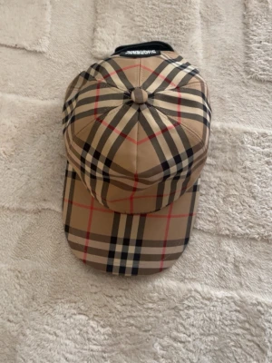 Beige Burberry keps  - Säljer en klassisk beige keps från Burberry med det ikoniska rutiga mönstret i svart, vitt och rött. Kepsen har justerbar rem bak och en patch med texten 'Burberry London England'. TPerfekt accessoar för en trendig och stilren look.