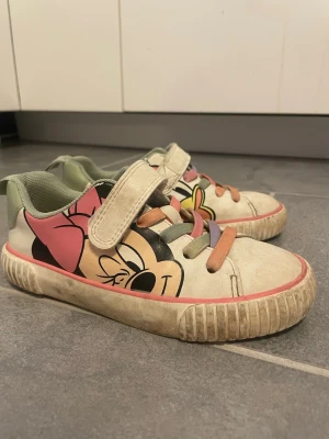 Sneakers med Minnie Mouse-tryck för barn - Snygga sneakers för barn med stort Minnie Mouse-tryck på sidan, vita med rosa och gröna detaljer. Skorna har kardborreband och elastiska snören i olika färger. Yttermaterialet är syntet och insidan är ljusgrön. Perfekta för dig som gillar Disney och vill sticka ut.