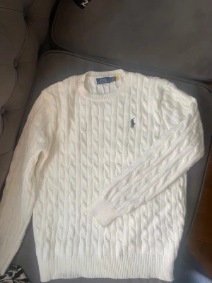 Vit kabelstickad tröja Polo Ralph Lauren - Kabelstickad vit tröja från Polo Ralph Lauren med rund halsringning och marinblå logga broderad på bröstet. Tröjan har ribbade muddar vid ärmslut och nederkant. Klassisk och tidlös design i mjukt material.