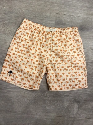 Beige badbyxor med palmtryck - Snygga badbyxor i beige med all-over tryck av bruna palmer och en liten svart palm broderad på benet. De har elastisk midja och är perfekta för stranden eller poolen. Materialet känns lätt och snabbtorkande.