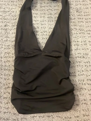 Svart halterneck topp med djup urringning - Snygg svart halterneck topp med djup v-ringning framtill och rynkade detaljer på sidorna. Toppens material är stretchigt och följsamt, perfekt för att skapa en cool och trendig look. Passar till både jeans och kjol för en stilren outfit.