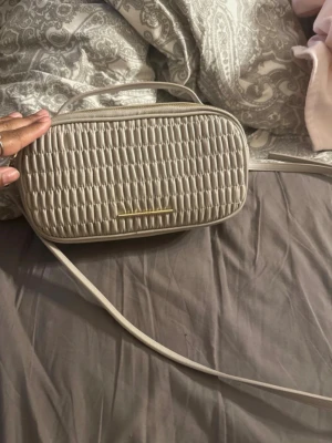 Beige oval Calvin Klein handväska  - Snygg beige handväska i oval form med flätad struktur på framsidan och guldfärgad detalj. Väskan har både axelrem och handtag, samt dragkedja upptill. Perfekt för dig som gillar stilrena accessoarer med en twist. Obs! Använd fåtal ggr o köpt för 1200kr