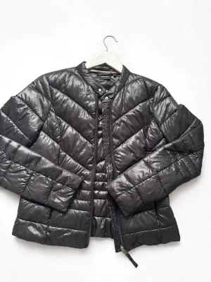Svart pufferjacka med rund krage - Snygg svart pufferjacka med rund krage och dragkedja framtill. Jackan har ett quiltat mönster i breda paneler och är tillverkad i ett glansigt material som ger en modern look. Perfekt för kalla dagar när du vill hålla värmen och se stilren ut. Märke H&M storlek 34
