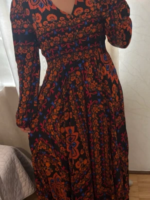 Färgstark långklänning med blommönster - Långklänning med intensivt blommönster i orange, blått och rött på svart botten. Klänningen har lång ärm, markerad midja och v-ringad hals. Tyget är mjukt och följsamt, perfekt för dig som gillar bohemiska vibes och färg.