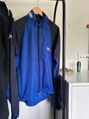 Blå och svart vindjacka med reflex - Snygg blå vindjacka med svarta ärmar och reflexdetaljer. Jackan har hel dragkedja framtill, hög krage och logga på bröstet. Tillverkad i lätt och vindtätt material, perfekt för blåsiga dagar. Praktisk ficka bak och ventilerande panel på ryggen. Ny pris runt 2500