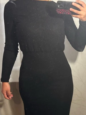 Svart glittrig långklänning  - Svart långklänning med diskret glittereffekt och lång ärm. Klänningen har en omlott-detalj i ryggen och markerad midja för en snygg siluett. Perfekt för dig som vill ha en elegant look med lite extra shine den är öppen framifrån ja hade den bara fel håll i bilden :) 