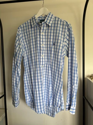 Blårutig skjorta från Ralph Lauren - Klassisk blårutig skjorta från Ralph Lauren med button-down krage och broderad logga på bröstet. Skjortan har långa ärmar, vita knappar och är tillverkad i mjuk bomull. Perfekt för en clean och stilren look. Passar som L