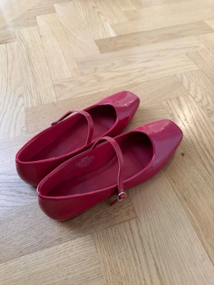 Röda lackade ballerinaskor med rem - Säljer ett par snygga ballerinaskor i blankt rosa lack med fyrkantig tå och justerbar rem över foten. Skorna har en klassisk Mary Jane-stil och ger en trendig touch till din outfit. Perfekta för dig som gillar färg och vill sticka ut. Endast använda en gång.