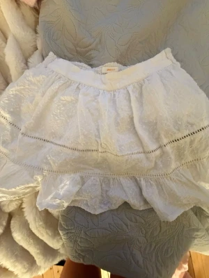 Vit volangkjol från Gina Tricot - Söt vit kjol från Gina Tricot med volanger och spetsdetaljer. Kjolen har elastisk midja och är gjord i ett lätt, luftigt tyg med diskret struktur. Perfekt för dig som gillar romantisk och feminin stil.