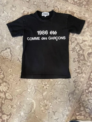 Svart CDG t-shirt - Svart t-shirt från CDG med vit text '1986 été' och logga på bröstet. Klassisk rund hals och korta ärmar. Perfekt för dig som gillar streetwear och vill ha något stilrent men ändå statement.