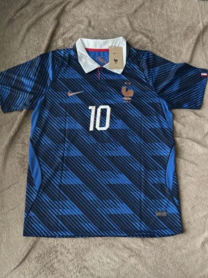 Frankrike fotbollströja Mbappé #10 - Snygg blå fotbollströja från Nike med Frankrikes landslagsemblem och nummer 10 på bröstet. Baksidan har namnet Mbappé och stort nummer 10. Tröjan har vit krage, korta ärmar och är tillverkad i lätt, ventilerande material.