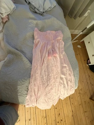 Ljusrosa långkjol från Gina Tricot - Söt ljusrosa lång kjolfrån Gina Tricot med smock upptill och dekorativa tofsar i midjan. Klänningen är axelbandslös och har en luftig, flowy passform som passar perfekt till varma dagar.