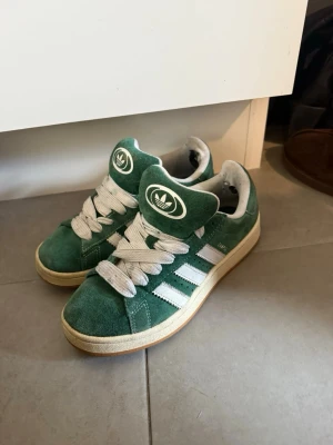 Gröna Adidas sneakers i mocka - Gröna Adidas sneakers i mocka med vita detaljer och klassiska tre ränder på sidan. Skorna har tjocka vita skosnören, gummisula i beige och Adidas-logga på plösen. Låg modell som passar perfekt till streetwear och vardagsstil.