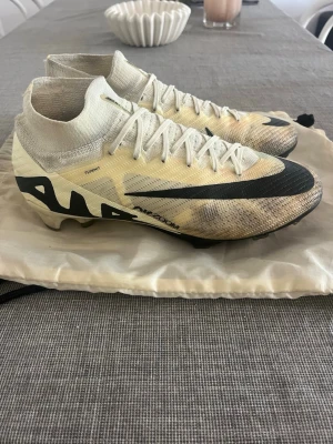 Nike Mercurial Superfly fotbollsskor - Säljer ett par Nike Mercurial Superfly fotbollsskor i beige och svart med högt skaft och snörning. Skorna har en strumpliknande passform och är tillverkade i ett lätt syntetmaterial för bästa bollkontroll och grepp på planen.