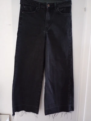Svarta vida jeans med fransig nederkant - Säljer ett par svarta jeans med hög midja och vida ben. Jeansen har råa, fransiga benslut och klassiska fem fickor. Tillverkade i denim med en cool, avslappnad look. Perfekta för dig som gillar trendiga och bekväma byxor.