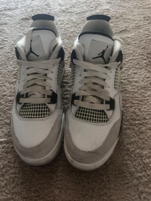 Vita och svarta Air Jordan 4 sneakers - Säljer ett par klassiska Air Jordan 4 sneakers i vitt och svart med mesh-detaljer på sidorna och tungan. Skorna har den ikoniska Jumpman-loggan både fram och bak samt svarta plastdetaljer vid snörningen. Perfekta för dig som gillar streetwear och sportig stil.