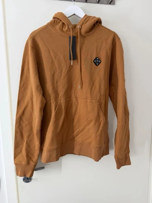 Brun hoodie med patch och ficka - Snygg brun hoodie med huva och dragsko samt en praktisk magficka. På bröstet sitter en liten svart patch. Perfekt för en avslappnad stil och enkel att matcha med jeans eller joggers!❤️