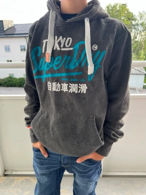 Mörkgrå Superdry hoodie med tryck - Säljer en mörkgrå hoodie från Superdry med stor turkos och vit logga samt japansk text på bröstet. Hoodien har vit dragsko, känguruficka och ribbade muddar. Storlek M.