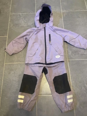 Ljuslila vindjacka med byxor  - Ljuslila vindbyxor och vindjacka med huva och dragkedja, svarta detaljer och en bröstficka med dragkedja. Jackan har två sidofickor och reflexdetaljer. Materialet är troligtvis polyester, perfekt för blåsiga dagar. Matchande byxor med svarta knäförstärkningar och reflexband vid benslut.