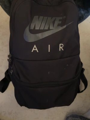Svart Nike Air ryggsäck - Snygg svart ryggsäck från Nike Air med stor logga framtill. Ryggsäcken har flera fack med dragkedja och justerbara axelband. Tillverkad i slitstarkt tyg, perfekt för skolan eller träningen. Enkel och stilren design som passar till det mesta.
