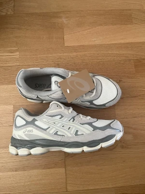Vita och grå sneakers från Asics - Snygga sneakers från Asics i vitt och ljusgrått med mesh- och mockadetaljer. Skorna har en chunky sula och klassisk Asics-logga på sidan. Perfekta för dig som gillar sportig och clean stil.
