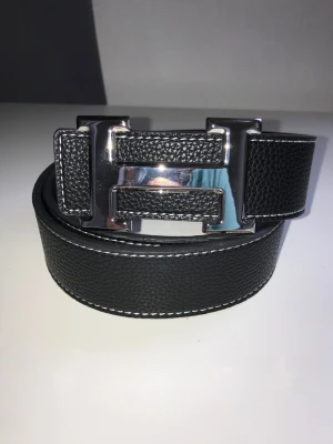 Svart läderbälte med stor H-spänne - Ny oanvänd Snyggt svart bälte i läder med vit kontrastsöm och ett stort, blankt H-format metallspänne. Bältet har en stilren och modern look som passar till jeans eller kostymbyxor. Perfekt accessoar för att lyfta din outfit.ett till ett,fraktar snabbt