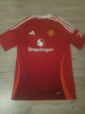 Manchester United matchtröja Adidas - Röd Manchester United matchtröja från Adidas med vita detaljer och klubbmärke på bröstet. Tröjan har Snapdragons logga på framsidan, vita ränder på axlarna och är tillverkad i ett lätt, ventilerande funktionsmaterial. Perfekt för fotbollsträning eller match. Den är helt ny.