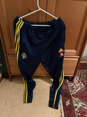 Blå adidas träningsbyxor Sverige - Mörkblå träningsbyxor från adidas med Sveriges landslagslogga och gula detaljer. Byxorna har tre gula ränder längs benen, resår i midjan och smal passform. Perfekta för fotbollsträning eller chill. Materialet är troligtvis polyester.