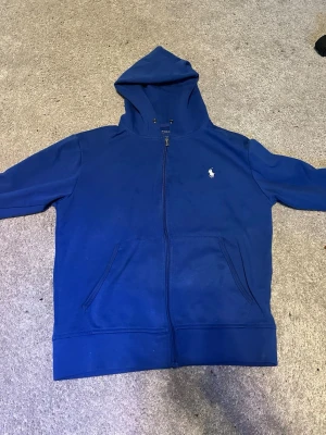 Blå zip hoodie från Polo Ralph Lauren - Snygg blå hoodie med dragkedja från Polo Ralph Lauren. Klassisk design med huva, två fickor framtill och den ikoniska vita logon broderad på bröstet. Perfekt för en chill och avslappnad stil. Tillverkad i mjukt bomullsmaterial.