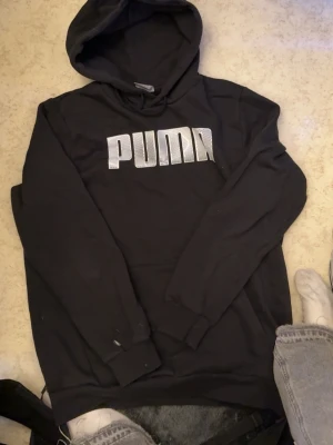 Svart hoodie från Puma med logga - Säljer en svart hoodie från Puma med stor vit logga på bröstet. Klassisk huva och magficka framtill. Tillverkad i mjukt material som känns skönt mot huden. Perfekt för dig som gillar sportig och avslappnad stil.
