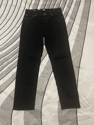 Svarta raka jeans - Säljer ett par svarta jeans med rak passform och klassisk femficksdesign. Jeansen har bälteshällor och knappgylf. Materialet ser ut att vara en bomullsblandning med lite stretch för extra komfort. Perfekta till en avslappnad stil.