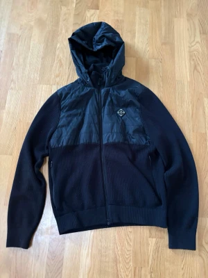 Svart cardigan med huva - Snygg svart cardigan med stickade ärmar och nederdel samt vadderad framsida och huva. Jackan har dragkedja framtill, två sidofickor och en liten patch på bröstet. 